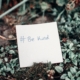 This picture shows a small garden, possibly a window garden or garden bed. Propped up in between some plans is a small square sign made of white paper that says #(hashtag) Be Kind